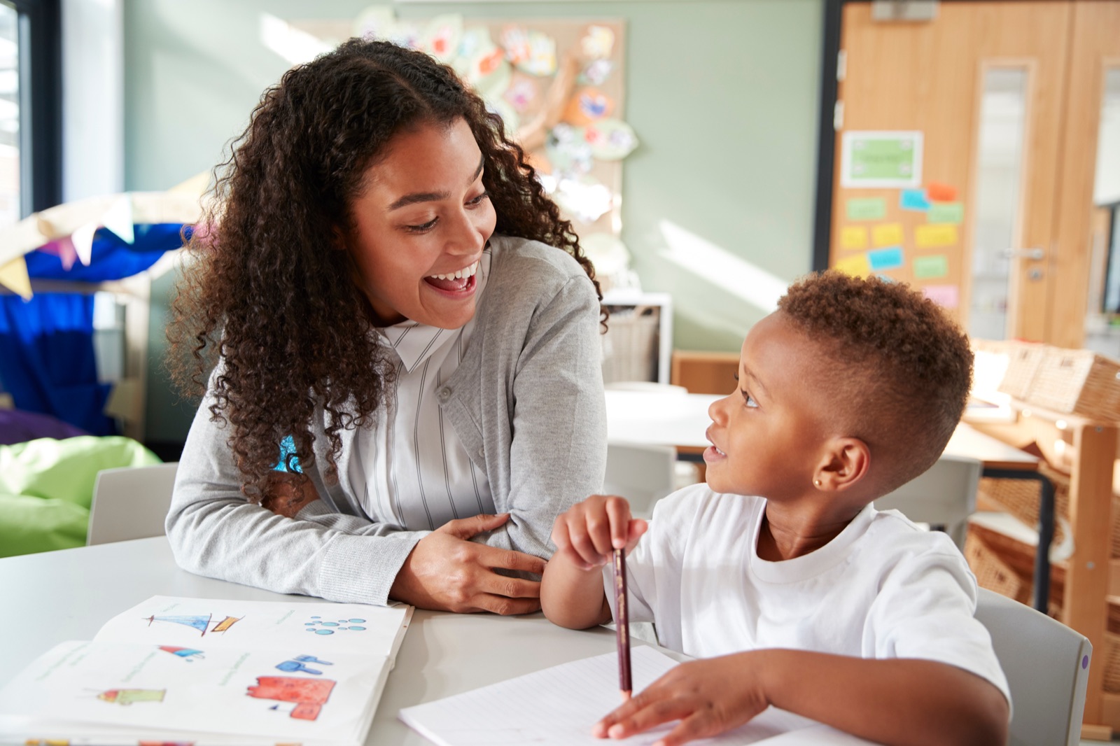 A teacher and a young student smile at each other while working together at a classroom table.